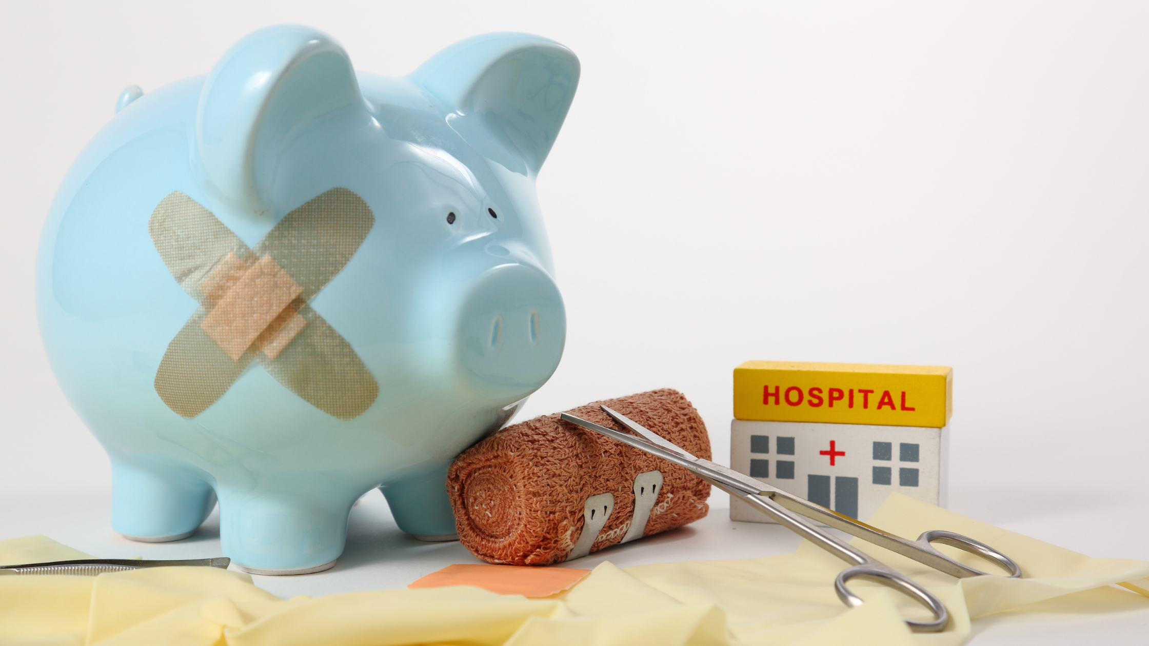 Blue piggy bank with band-aids on its' side. A roll of gauze, surgical scissors, and a small block shaped like a hospital are next to it.