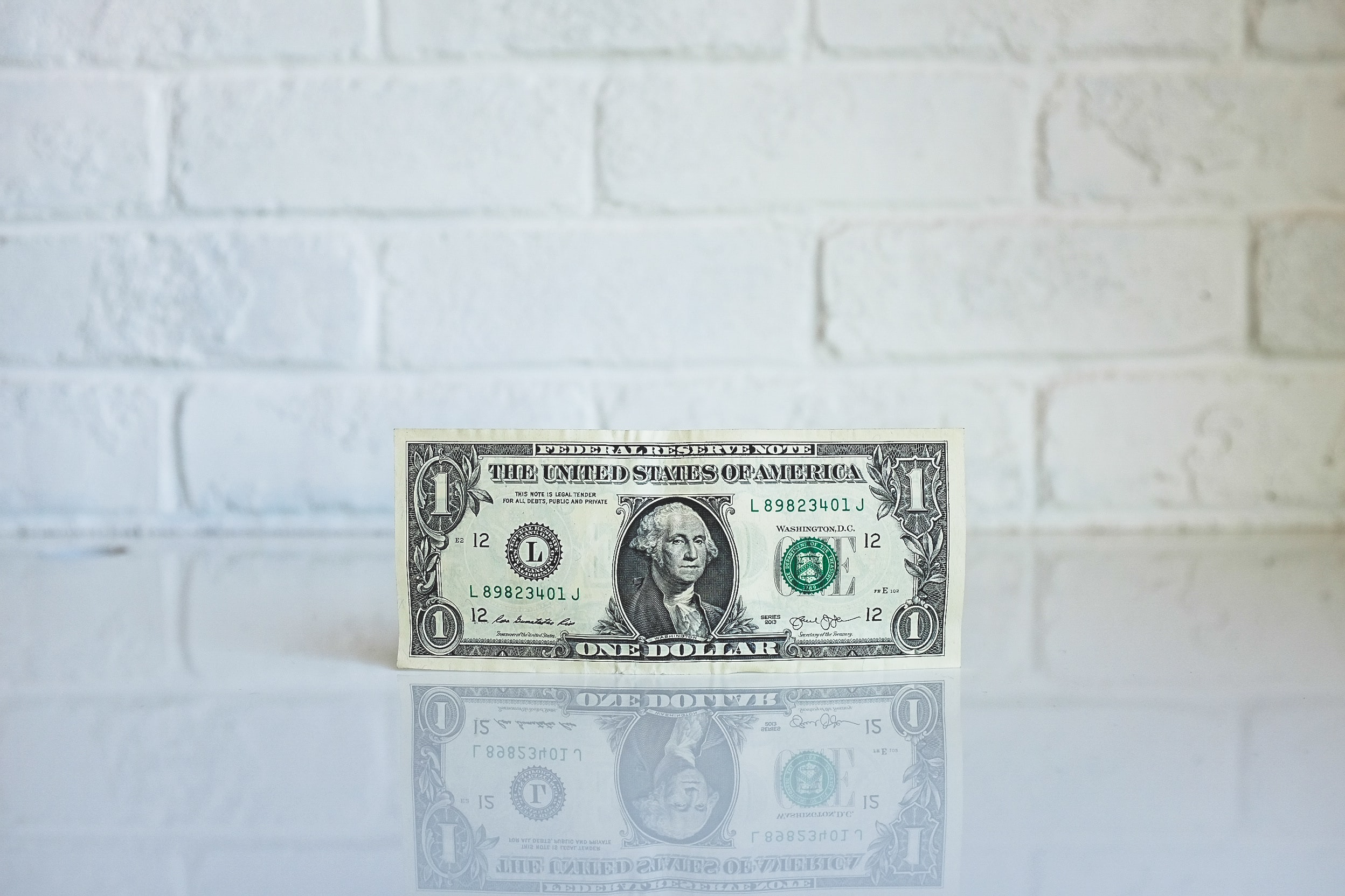 Image of a one dollar bill against a white brick background. Photo by NeONBRAND on Unsplash.